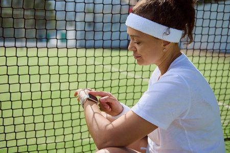 A female athlete sitting near a tennis net checks her smartwatch for performance data. She wears a white sports outfit, conveying determination and modern technology in sports.の写真素材