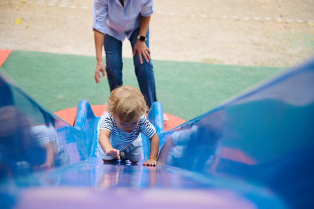 A young child climbs up a blue slide in a lively playground setting while a parent provides...
