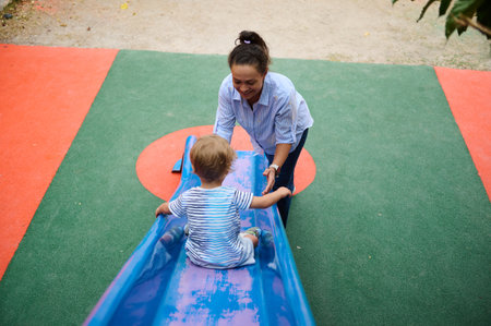 A caring mother assists her young child as they play on a slide at a vibrant outdoor playground,...