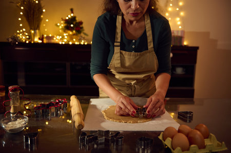 A woman in an apron is cutting cookie dough into festive shapes in a cozy, Christmas-lit kitchen....