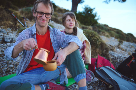 A couple relaxes on rocky terrain during an outdoor adventure, brewing a drink using a copper kettle. Surrounded by backpacks and the beauty of nature, they enjoy the serenity of camping in the countryside.の写真素材