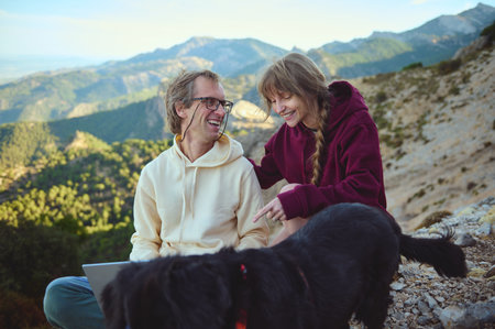A happy couple relaxes on a mountain trail with their black dog and a laptop. They enjoy the serene natural surroundings, showcasing enjoyment, bonding, and a break from daily routines.の写真素材