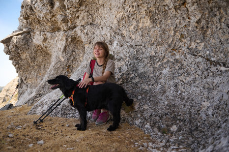 A young woman enjoying a peaceful moment on a hike with her black dog by her side, sitting near a textured rock formation under a sunny sky. The scene exudes tranquility and companionship.の写真素材