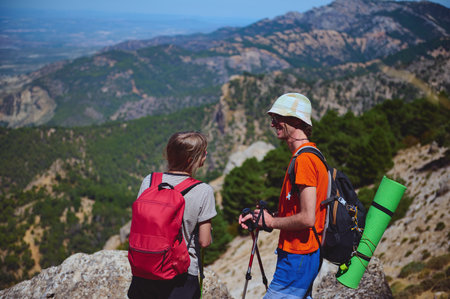 Two hikers with backpacks stand atop a mountain, appreciating the breathtaking natural scenery. The sunny weather and green surroundings create a sense of joy and adventure in this outdoor activity.の写真素材
