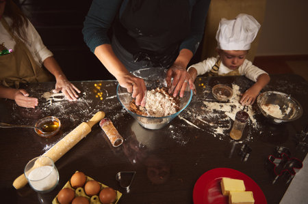 A warm scene of a mother and two children cooking and baking gingerbread at a kitchen table, flour...