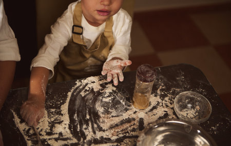 A young child with flour on hands leans over a dark wooden table, surrounded by bowls and jars as...