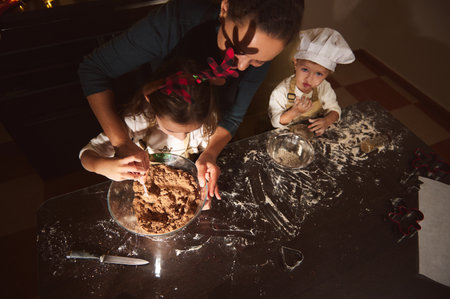 A happy mother guides two children in gingerbread making, flour scattered on a dark table, festive...