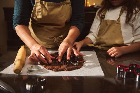 A mother and child in aprons press cookie cutters into brown gingerbread dough on parchment, with a...
