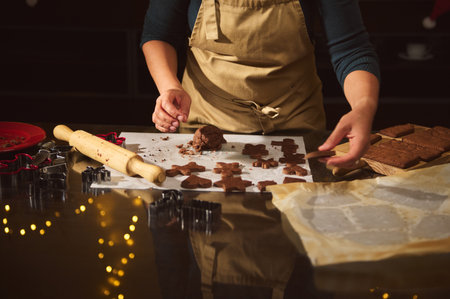 A person in a beige apron shapes gingerbread cookies on parchment, with a rolling pin and cutters...
