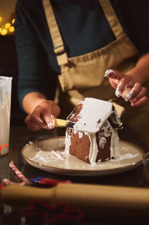 A cozy kitchen scene shows a baker decorating a gingerbread house with white icing, capturing warm...