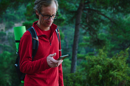 A man in a red fleece checks his smartphone on a forest trail, carrying a backpack with a green...