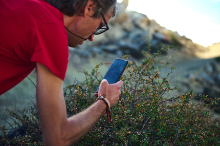 A man in a red shirt leans toward a smartphone among shrubs in a rocky outdoor setting, conveying...