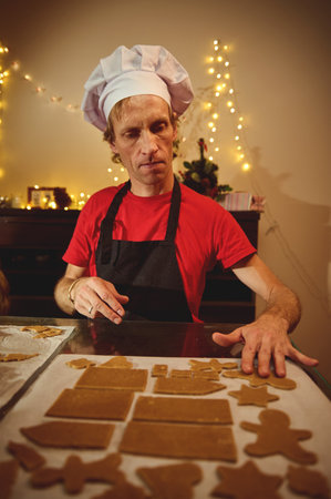 Focused baker in a chef hat and red shirt shapes gingerbread cookies on parchment, as warm Christmas...