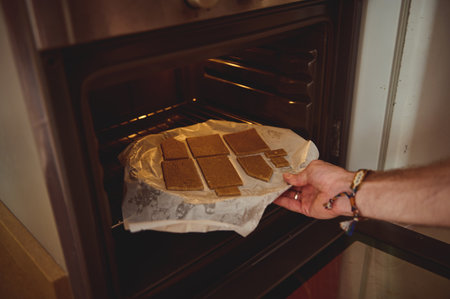 A hand places a parchment-lined tray of brownie bars into an open oven, creating a warm, cozy...
