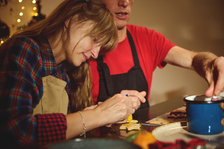Two adults decorate cookies in a warm, festive kitchen. A man in red with an apron helps while a woman smiles, creating a cozy Christmas moment of family, friends, and baking.の写真素材