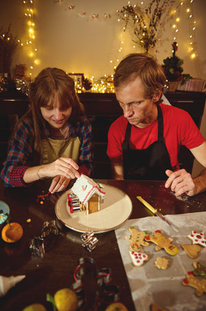 Two people in aprons decorate a gingerbread house while cookies cool nearby, illuminated by string...