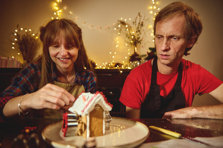 A man and woman in aprons decorate a gingerbread house at a warmly lit holiday table, smiling as...