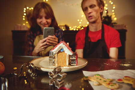 A warm Christmas kitchen scene with a gingerbread house centerpiece, twinkling lights, and a smiling couple filming the moment on a phone.の写真素材