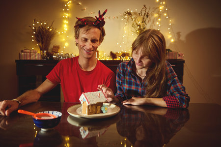 Smiling couple at a dining table decorates a gingerbread house amid warm fairy lights, capturing...
