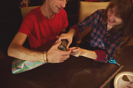 A couple laughs together while one person helps unwrap a colorful wrapped present at a wooden table, capturing a warm, festive home moment.の写真素材