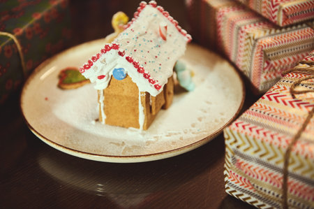 Gingerbread house sits on a plate, dusted with sugar, amid wrapped presents for a cozy Christmas indoor scene, suggesting family holiday baking and gift-giving warmth.の写真素材