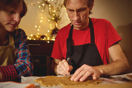 Two adults bake in a warm kitchen, man in a red shirt and apron slices dough while a woman watches, surrounded by festive Christmas lights.の写真素材