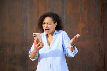 A surprised woman in a blue striped shirt looks at her phone with an open mouth and raised hand,...