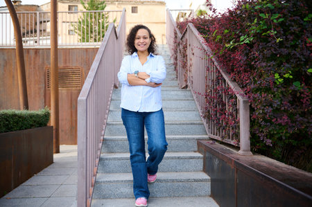 A smiling woman in a light blue shirt and jeans walks down a modern outdoor staircase, arms crossed....