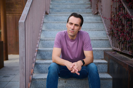 A man in a purple t-shirt sits on outdoor steps, arms resting, deep in thought. Urban casual...