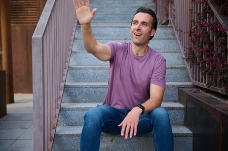 A joyful man sits on outdoor stairs, waving with a smile, wearing a purple tee and jeans for a...