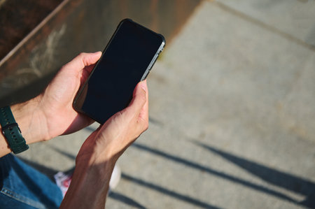 Close-up of hands holding a smartphone outdoors on a sunny day. The device rests in a...