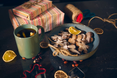 A warm Christmas table with a green mug of tea, a blue plate of sugar-dusted cookies, wrapped gifts, and lemon slices for festive charm and homey delight.の写真素材