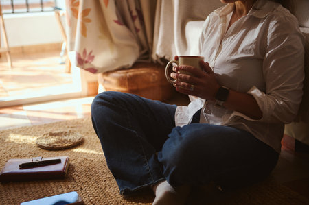 A woman sits cross-legged on a woven rug, enjoying coffee in a sunlit, cozy living corner. Casual shirt, jeans, smartwatch, notebook, and phone nearby.の写真素材