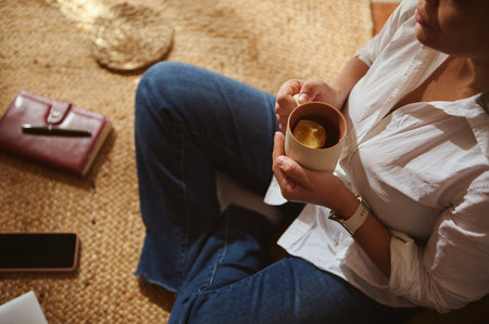 A woman sits on a woven rug, wearing jeans and a white shirt, enjoying tea from a mug with lemon. Calm, casual, cozy home moment.の写真素材