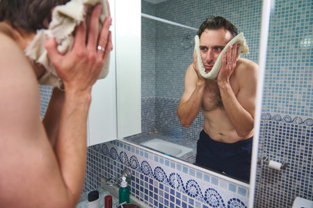 A shirtless man stands at a bathroom sink, wiping his face with a towel while looking into a mirror. Blue mosaic tiles create a calm setting, emphasizing daily grooming and self-care.の写真素材