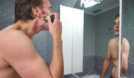 Man shaves in a tiled bathroom, applying cream with a brush and razor, seen in the mirror. Focus on grooming, daily routine, self-care.の写真素材