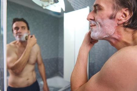 A shirtless man stands in a modern bathroom, applying shaving foam and preparing to shave. Reflected in the mirror, he appears focused and relaxed, capturing a daily grooming moment.の写真素材