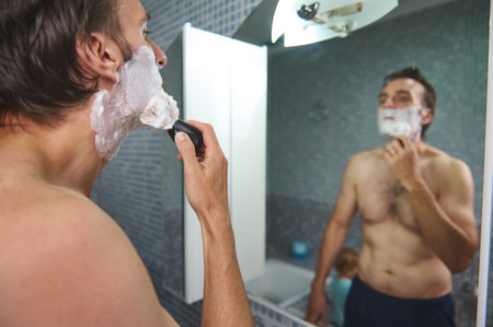 A man shaves in a bright bathroom, applying foam and using a razor in the mirror. A child watches nearby, creating a casual, intimate domestic moment of grooming and family life.の写真素材