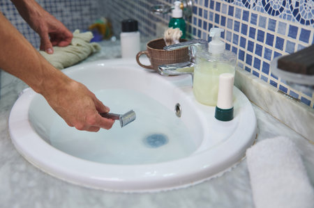 A close-up view of a person shaving at a bathroom sink. Faucet, soap dispenser, and grooming items rest on the marble countertop, with cool blue tiled wall in background.の写真素材