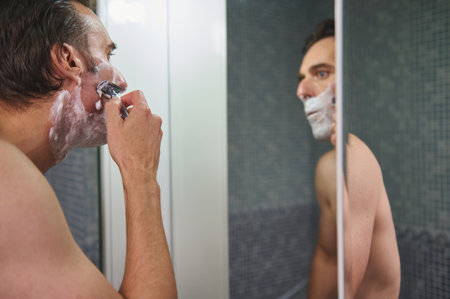 A shirtless man lathers foam on his face while shaving in a tiled bathroom, reflected in the mirror. A calm moment of daily grooming and self-care.の写真素材