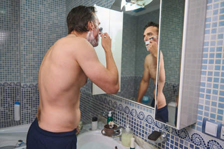 A shirtless man stands at a bathroom sink, applying shaving foam and checking his reflection in the mirror, capturing a personal care routine.の写真素材