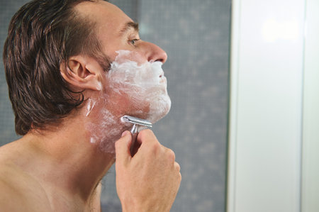 A man shaves in a bathroom, applying foam and using a razor. Side profile captures grooming routine, clean skin, and a focused, everyday moment.の写真素材