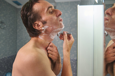 A man stands shirtless before a bathroom mirror, shaving with a razor. Soap lathers on his chin as a hand guides his neck. A moment of grooming and daily routine.の写真素材