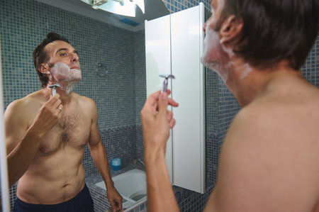 A shirtless man shaves in a tiled bathroom, foam on his face, captured in a reflective mirror for a personal care moment.の写真素材