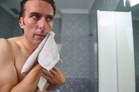 Casual, shirtless man in a modern bathroom uses a white towel to dry his face, conveying self-care and a daily morning routine.の写真素材
