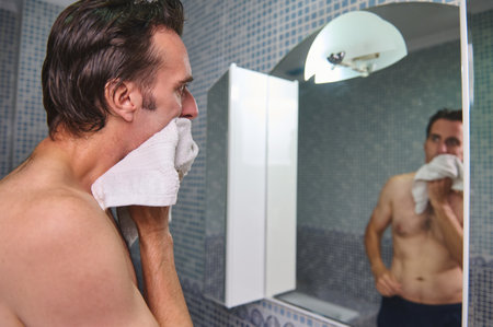 A shirtless man dries his face with a towel in a tiled bathroom, seen from the side with his reflection in the mirror. The scene conveys routine grooming and calm morning moments.の写真素材