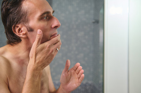 A man stands in a modern bathroom, deep in thought with his hand against his chin. The intimate close-up captures contemplation, privacy, and everyday morning routines.の写真素材