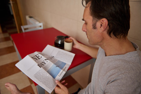 A man sits at a red table in a cozy kitchen, reading a newspaper while enjoying a cup of coffee. A calm, everyday morning moment captured candidly.の写真素材