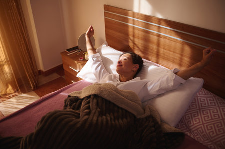 A woman lies in bed, arms stretched, enjoying warm sunrise light. White shirt, soft blanket, cozy bedroom setting conveying relaxation, wellness, morning routine, and home comfort.の写真素材