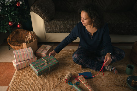 A woman sits on a woven rug near a Christmas tree, wrapping presents with ribbon and tape. Warm, festive scene conveys home, family, and holiday preparation.の写真素材